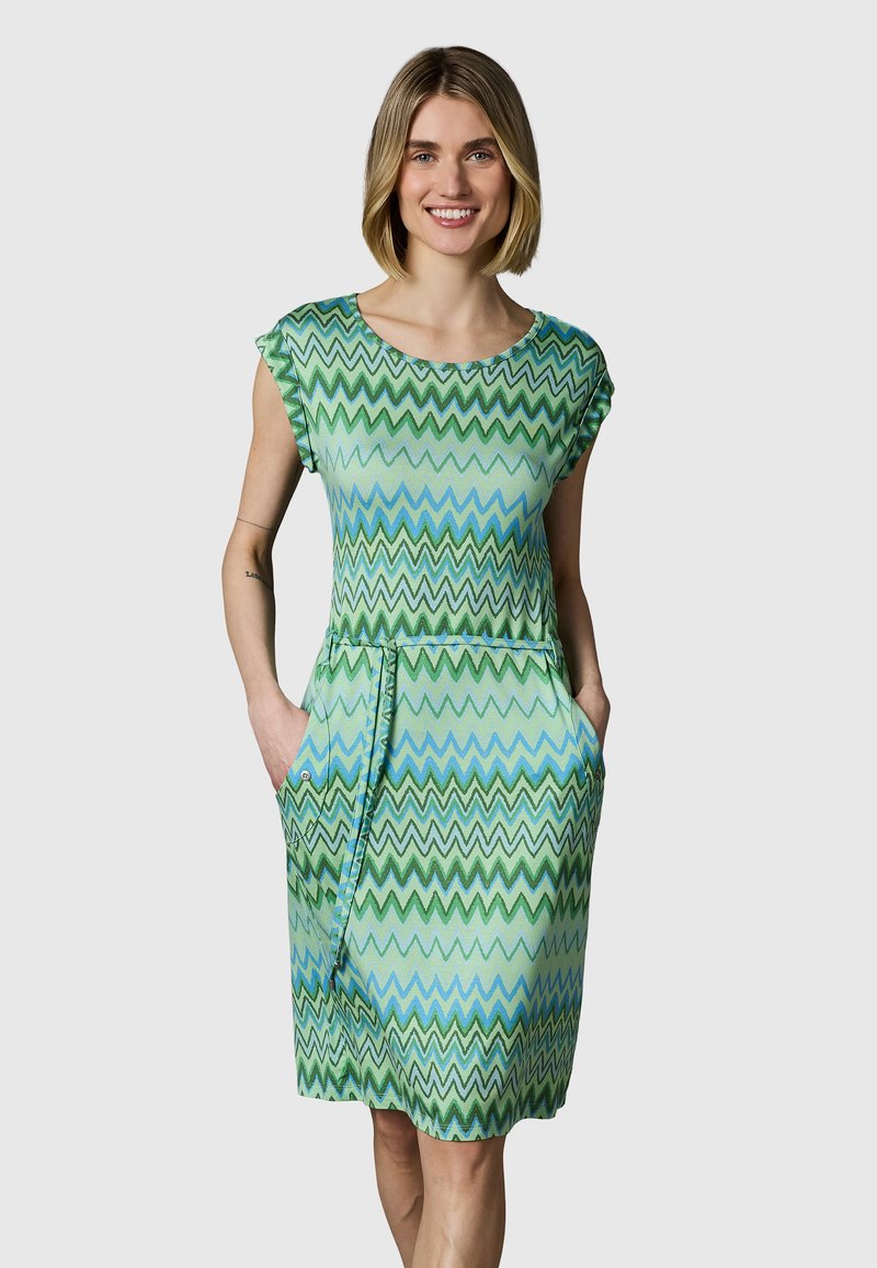 Vrouw draagt een groene en blauwe zigzagpatroon knielange jurk met korte mouwen en twee voorzakken, glimlachend met handen in de zakken.