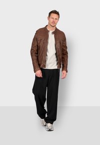Giacca di pelle marrone con due tasche con zip, abbinata a una t-shirt bianca e pantaloni neri larghi, completata da sneakers grigie.