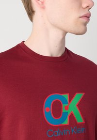 Burgundy színű pamut póló, amelyen egy színes "OK" logó található kék és zöld színekben, piros kiegészítőkkel, alatta a "Calvin Klein" márkanevével.