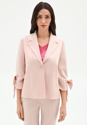 Donna con lunghi capelli scuri che indossa una giacca rosa pallido con maniche 3/4 annodate sopra una maglietta rosa con bordo in pizzo e pantaloni rosa pallido abbinati.