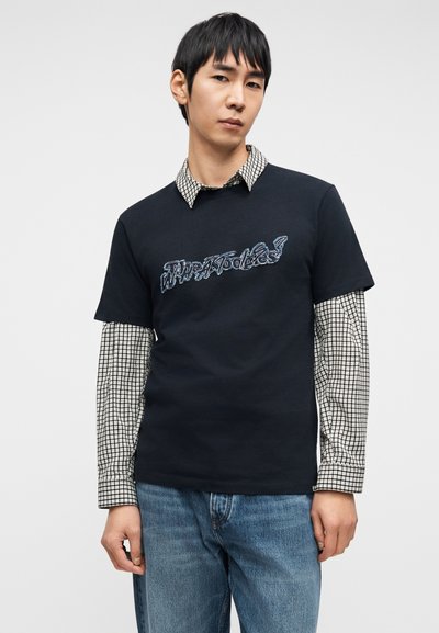 Μαύρο T-shirt με κεντημένο κείμενο, φορεμένο πάνω από λευκό και μαύρο καρό μακρυμάνικο πουκάμισο. Συνδυάζεται με τζιν παντελόνι ανοιχτού γαλάζιου χρώματος.