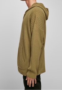Oliwkowa zielona prążkowana bluza z kapturem o luźnym kroju, teksturowanym materiale, kieszenią kangurka i wyraźnym kapturem. Posiada długie rękawy i casualowy fason.