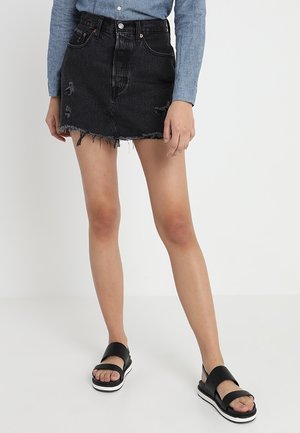 Kobieta ubrana w czarną, postrzępioną mini spódnicę z denimu, niebieską koszulę z długim rękawem i czarne sandały na platformie, stojąca na białym tle.