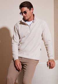 Svijetlosmeđi zip-up flis s visokim ovratnikom i rebrastim manžetama, u paru sa svijetlosmeđim hlačama. Glatka tekstura, ležeran kroj.