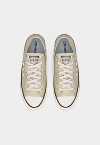 Ζευγάρι μπεζ χαμηλά αθλητικά παπούτσια Converse All Star με λευκά κορδόνια και διακοσμητικές λευκές λεπτομέρειες σε σχήμα λουλουδιών στα πλευρά.