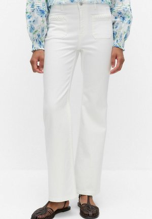 Pantalon blanc droit avec poches plaquées à l'avant et bordure tressée, porté avec des sandales plates noires tissées et un chemisier bleu à motifs floraux.