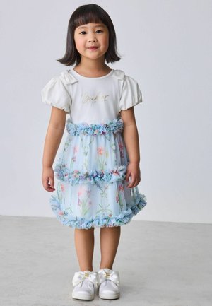 Ung pige med kort sort hår iført en hvid bluse med pufærmer, blå blomsterprintet flæsekjol og hvide sko med store sløjfer.