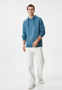 Sudadera azul hecha de tejido suave, con un bolsillo canguro, cordones y puños de canalé, combinada con pantalones cargo blancos y zapatillas.
