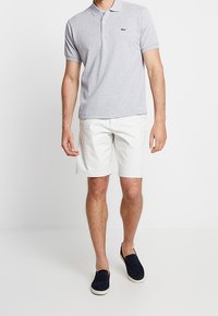 Hombre vestido con una camiseta polo gris claro, pantalones cortos blancos y zapatos de lona oscuros, de pie contra un fondo blanco plano.