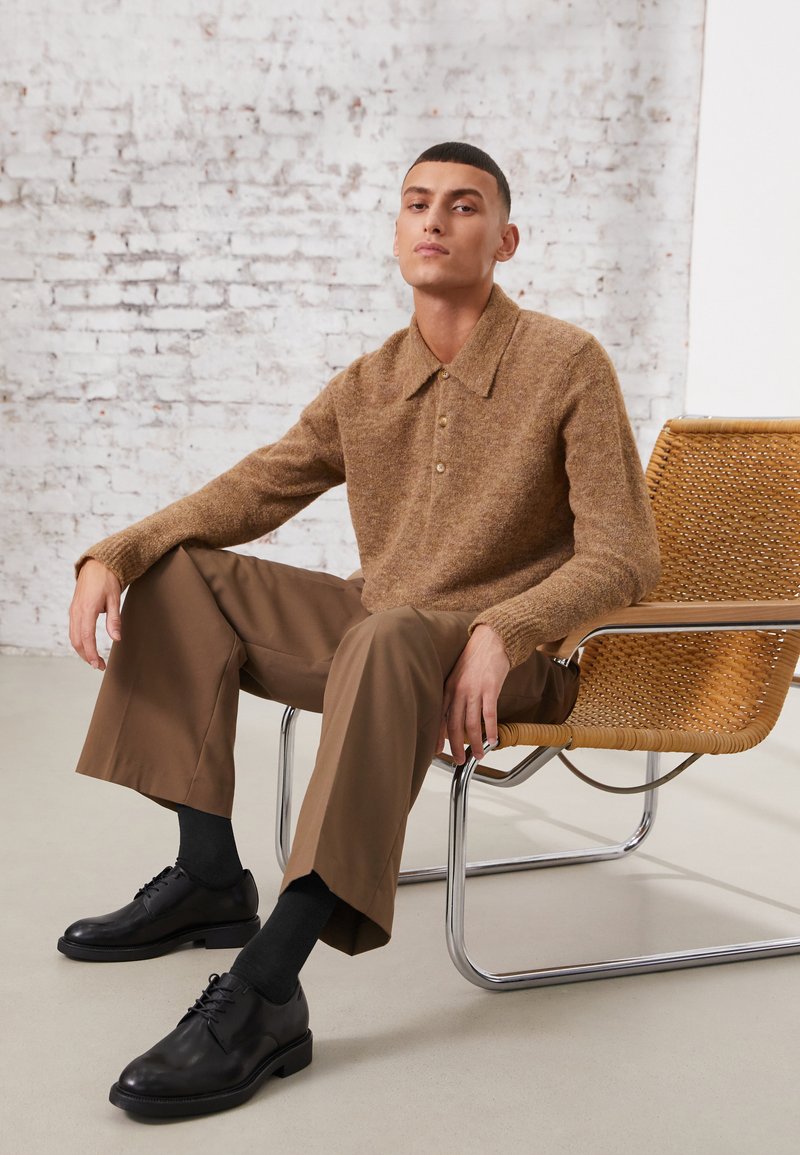 Polo di maglia marrone in lana con patta bottonata, indossato con pantaloni larghi marroni e scarpe in pelle nere. Sedia intrecciata sullo sfondo.