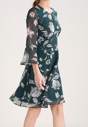 Kobieta w ciemnozielonej sukience do kolan z białymi kwiatowymi wzorami i przejrzystymi, trzy czwarte rękawami w kształcie dzwonu, stojąca na jednolitym tle.