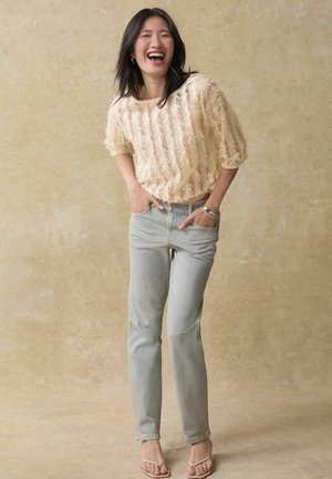 Haut en tricot texturé beige clair avec des manches courtes, associé à un jean droit bleu clair et des sandales claires. Design simple et couleurs neutres.