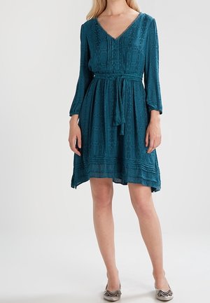 Femme portant une robe bleu sarcelle longueur genou avec manches longues, col en V et ceinture à la taille, associée à des chaussures plates beiges sur fond blanc.