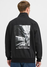 Sudadera negra con un cuello alto y una gran impresión gráfica en la parte posterior. La impresión representa una escena de metro con texto debajo.
