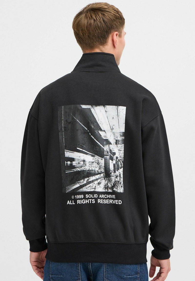 Sudadera negra con un cuello alto y una gran impresión gráfica en la parte posterior. La impresión representa una escena de metro con texto debajo.