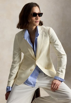 Frau mit schulterlangem braunem Haar trägt dunkle Sonnenbrille, cremefarbenen Blazer, hellblaues Hemd und weiße Hose, sitzt vor neutralem Hintergrund.