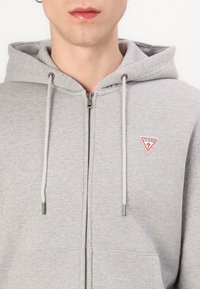 Felpa grigia con zip realizzata in tessuto morbido, caratterizzata da una tasca a marsupio, cappuccio con cordino e un patch a forma di triangolo rosso sul petto.