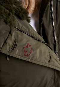 Olivově zelená puffa bunda s límcem z umělé kožešiny, s červeným vyšitým motivem listu a kapsami na zip. Viditelná strukturovaná tkanina a švy.