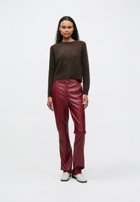 Una donna è in piedi contro uno sfondo bianco indossando un maglione marrone scuro, pantaloni in pelle bordeaux con cerniera frontale e scarpe slip-on bianche.