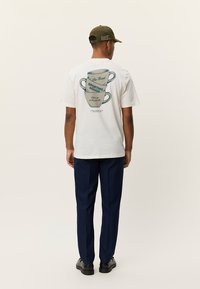 Λευκό κοντομάνικο T-shirt με μια εικόνα δύο κούπες στην πλάτη, με μπλε κείμενο. Στολή ολοκληρώνεται με σκούρο μπλε παντελόνι και μαύρα παπούτσια.