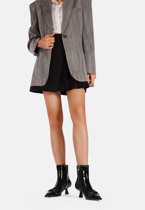 Blazer grigio in suede sopra una camicetta bianca, abbinato a una minigonna nera. Stivaletti neri in vernice con punta a mandorla e dettagli decorativi.