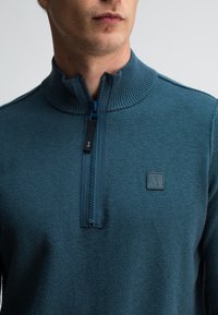 Türkiser Reißverschluss-Pullover mit hohem Kragen, gerippter Textur und einem kleinen Logo-Patch in der Nähe des Reißverschlusses. Verfügt über einen kontrastierenden blauen Reißverschluss.