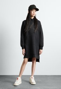 Čierne mikinové šaty s kangaroo vreckom a zlatým logom, s rebrovanými manžetami a spodným okrajom. Skombinované s béžovými teniskami a čiernou čiapkou.
