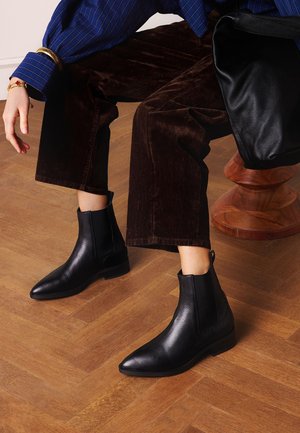 Person trägt schwarze Leder-Stiefeletten, braune Cordhosen und ein blau gestreiftes Hemd, sitzt auf einem Holzstuhl und hält eine schwarze Lederhandtasche.