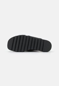 Suola di scarpa nera con profonde scanalature orizzontali e tallone texturizzato, vista dal basso su uno sfondo bianco uniforme.