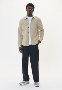Camicia beige con bottoni e due tasche sul petto, indossata sopra una maglietta bianca. Abbinata a pantaloni neri e scarpe sportive grigie. Sfondo chiaro.