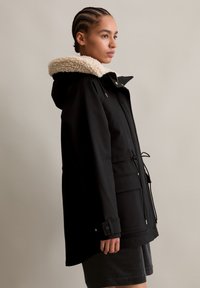 Zwarte parka met een crème sherpa-gevoerde capuchon, verstelbare trekkoord in de taille en voorzakken. Gladde textuur en contrasterende kleurdetails.