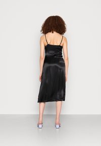 Vestido midi de cetim preto com alças finas, apresentando uma textura suave e um corte reto, alcançando a metade da panturrilha. Com sandálias de salto roxas.