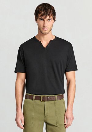 Uomo con maglia henley nera a maniche corte infilata nei pantaloni verde oliva con cintura marrone, in piedi davanti a uno sfondo semplice.