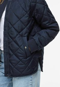 Temno modra prešita bomber jakna z diagonalnim šivanjem, elastičnimi manšetami in stranskim žepom z metalnim pritisnim gumbom. Gladek, lahek material.