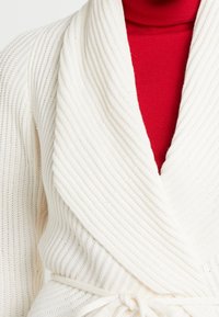 Cárdigan envuelto de punto acanalado blanco con un amplio cuello y cinturilla anudada, superpuesto a un cuello alto rojo. Presenta un patrón texturizado.
