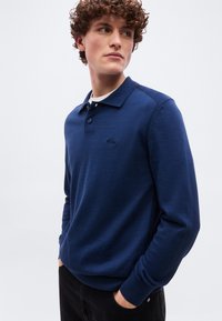 Νέος άνδρας με σγουρά μαλλιά, φορώντας σκούρο μπλε πόλο με μακριά μανίκια, με τα χέρια στις τσέπες, κοιτάζοντας ελαφρώς προς το πλάι μπροστά σε απλό φόντο.