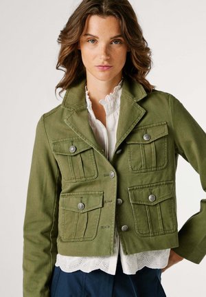 Kobieta o falujących brązowych włosach, ubrana w oliwkowozieloną, krótką kurtkę z czterema kieszeniami na guziki, na białej, falbaniastej bluzce i granatowych spodniach.