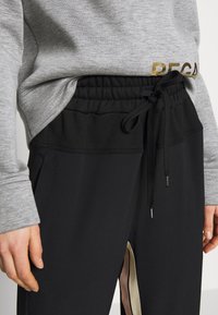 Svarta träningsbyxor med dragsko i midjan, med en sidoklyfta accentuerad med en ljusbeige panel. Grå långärmad topp med guldtext.