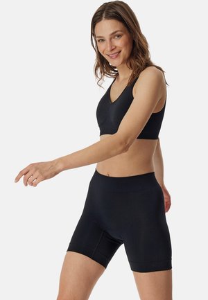 Lächelnde Frau mit schulterlangem braunem Haar, die einen schwarzen Sport-BH und eng anliegende Radlerhosen vor einfachem Hintergrund trägt.