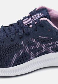 Navy blue urheilujalkine, jossa on verkkomateriaali, vaaleanpunaiset yksityiskohdat, teksturoitu pohja ja logo kielekkeessä. Varustettu nauhakiristyksellä ja kuvioidulla sivusuunnittelulla.