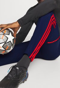 Laivaston siniset urheiluhousut, joissa punaiset raidat, yhdistettynä mustaan pitkähihainen paita. Kädessä pidetään mustaa jalkapalloa, jossa on oransseja yksityiskohtia. Mustat jalkapallokengät.