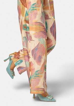 Mujer que lleva pantalones anchos con estampado de plumas en tonos pastel y tacones altos de ante verde azulado adornados con hebillas con joyas.