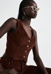 Bruine corduroy gecropped vest met een V-hals en vijf gouden knopen. Model draagt lichtgekleurde retro-stijl zonnebril en eenvoudige oorbellen.