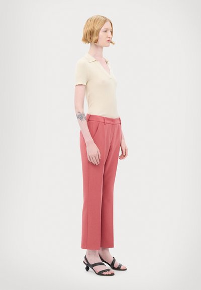 Polo beige à côtes avec un col en V, associé à un pantalon rose bien coupé. Sandales noires à glisser avec un accent rond. Fond plat.