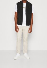 Schwarze ärmellose Weste über einem weißen T-Shirt, kombiniert mit hellbeigen Hosen und weißen Sneakers. Glatte Texturen und schlichtes Design.