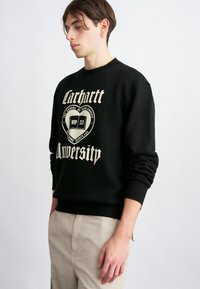 Czarna bluza z białym tekstem graficznym „Carhartt University”. Okr�ągły dekolt, ściągacze przy rękawach i u dołu, prezentująca miękką teksturę materiału.