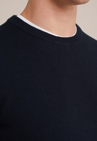 Man som bär en svart strukturerad stickad tröja över en vit T-shirt med rund halsringning, visar hals och nedre del av ansiktet mot en beige bakgrund.