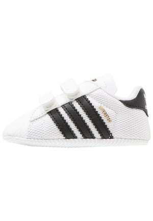 Hvid Adidas Superstar sneaker med sorte striber, velcrolukninger og guldfarvet "SUPERSTAR" tekst på en tekstureret stofoverflade.