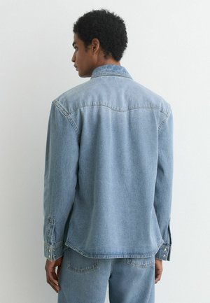 Mand set fra siden iført lyseblå denimskjorte og matchende jeans foran en ensfarvet hvid baggrund.