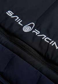 Svart isolerad jacka med kontrasterande marinblått tyg, med en synlig dragkedja och en vit broderad "Sail Racing"-logotyp.